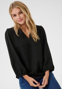 Black V-neck blouse in textured fabric, featuring long puff sleeves with elastic cuffs, paired with blue jeans, model's hands are visible.