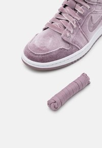 Detailní záběr na fialové semišové tenisky s bílou podrážkou a odpovídajícími stočenými tkaničkami na bílém pozadí.