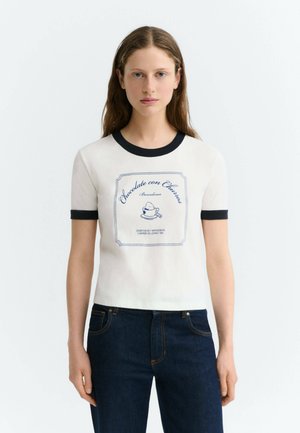 Femme aux longs cheveux bruns portant un t-shirt blanc à manches courtes avec bordure noire et texte et motif bleus, associée à un jean bleu foncé.