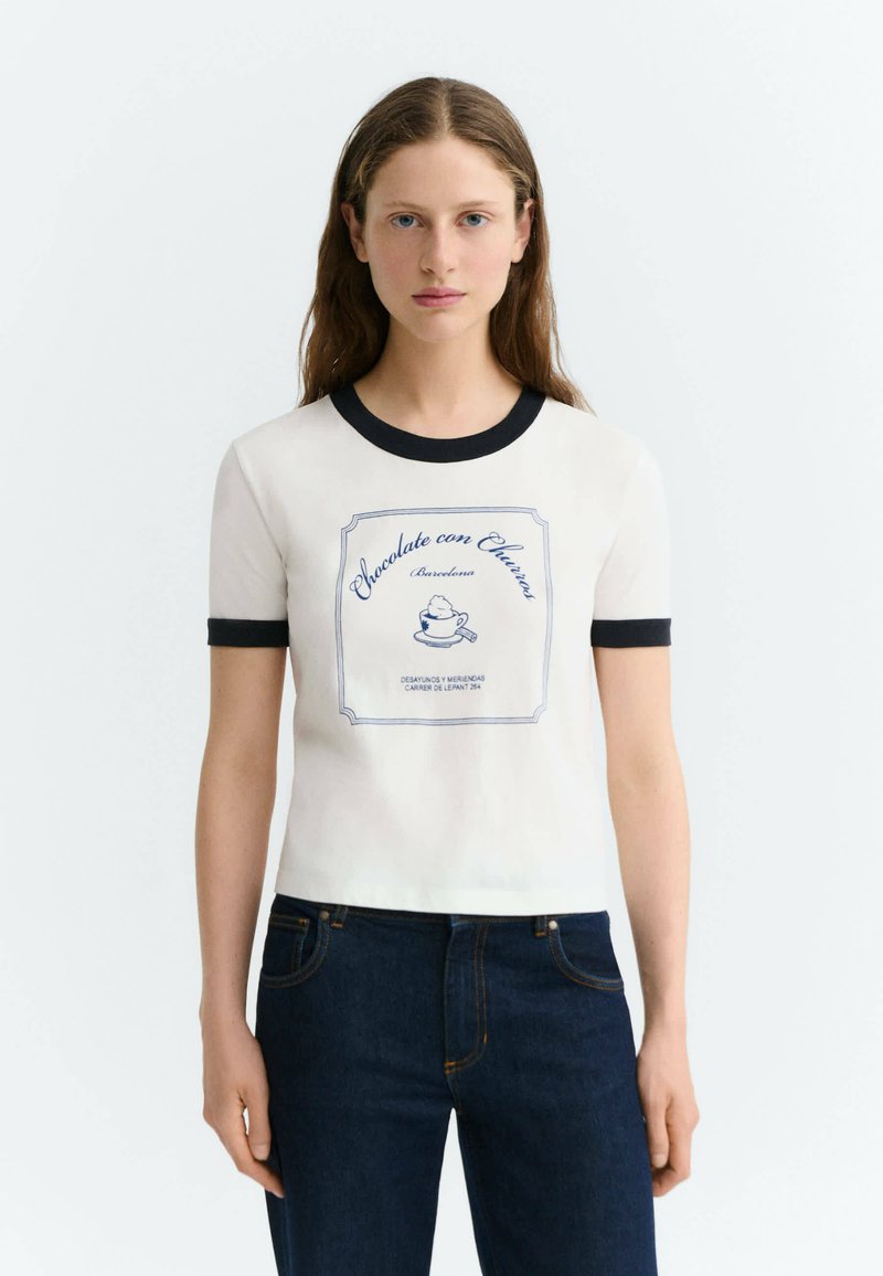 Femme aux longs cheveux bruns portant un t-shirt blanc à manches courtes avec bordure noire et texte et motif bleus, associée à un jean bleu foncé.