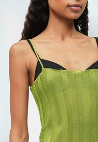 Zelený žebrovaný top s tenkými ramínky, s texturovaným povrchem a přiléhavým střihem. Vnitřní vrstva je černá, což vytváří vrstvený vzhled.