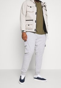 Muž v světle béžové bundě s kapucí, olivovém tričku, světle šedých kapsáčích a modro-bílých teniskách stojí u bílé zdi.