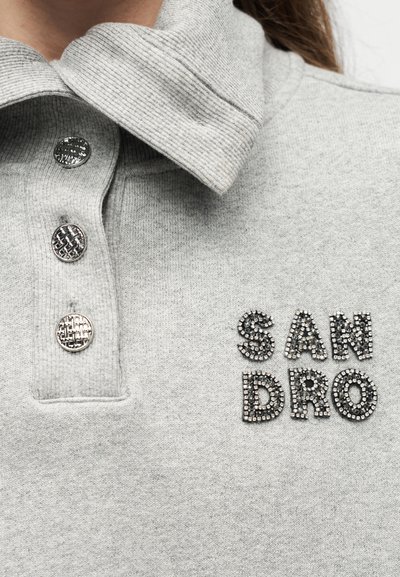 Közelkép egy szürke galléros pulóverről, ezüstös textúrájú gombokkal és fekete, gyöngyökkel díszített "SAN DRO" felirattal a mellrészen.