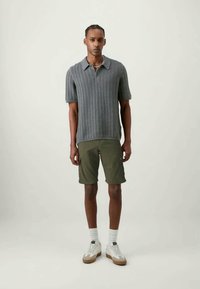 Camisola polo cinza tricotada com colarinho, combinada com calções verde-azeitona. Usada com ténis brancos com sola castanha e meias brancas.