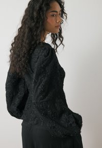 Musta tekstuuri blouse, jossa on kukkakuviot, puffihuput ja rento istuvuus. Kangas esittelee ryppyisen muotoilun lisäulottuvuuden vuoksi.