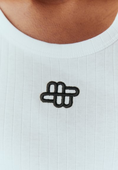 Haut en tissu blanc côtelé avec un logo noir brodé représentant des lignes entrelacées au niveau de la poitrine. Détail de col rond simple.
