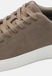 Brune skinnsneaker med lav profil, med teksturert overdel, flate snorer og hvit gummisåle. Synlig sømdetalj.