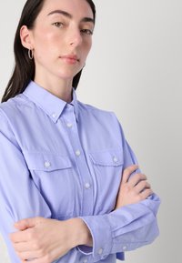 Camicia button-up di un leggero colore viola con due tasche sul petto, tessuto morbido, maniche lunghe e colletto classico. Presenta chiusura a bottoni e polsini.