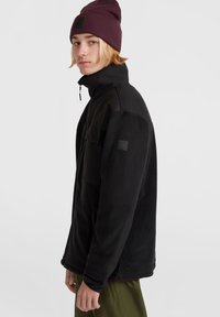 Giacca in pile nera con colletto alto, chiusura a zip, tasche laterali e pannello in tessuto a contrasto sulle spalle; indossata con un berretto bordeaux.