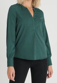 Grön blus med rutigt mönster, långa ärmar, V-ringning och samlad manschettdetalj. Lättviktsmaterial med en mjuk textur.
