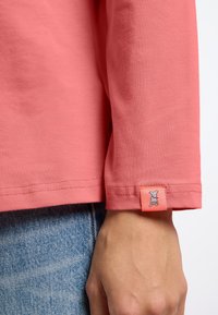 Koronkowa koszulka z długim rękawem w kolorze koralowym, o miękkiej fakturze, z pomarańczową metką z haftowanym wzorem psa przy mankiecie. Widać dżinsy.