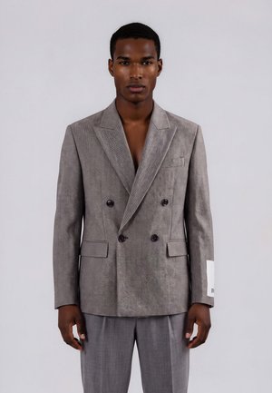 Man draagt een grijs dubbelgeknoopte blazer met zwarte knopen en bijpassende grijze broek, staand tegen een eenvoudige lichte achtergrond.