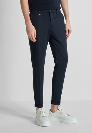 Hombre con pantalones ajustados azul marino oscuro remangados en los puños y zapatillas blancas con cordones, de pie sobre un suelo y fondo gris claro.