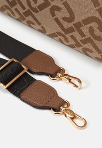 Tracolla regolabile in tessuto nero con estremità in pelle marrone testurizzata e fibbie metalliche dorate accanto a una borsa in tessuto marrone a motivo.