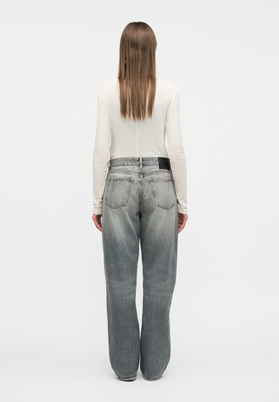 Γυναίκα με μακριά ίσια μαλλιά που φοράει εφαρμοστό λευκό μακρυμάνικο τοπ και φαρδιά, ξεθωριασμένα μπλε τζιν, στέκεται με την πλάτη προς τα εμπρός σε απλό φόντο.