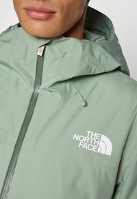 Chaqueta con capucha de color verde claro hecha de un tejido suave, que presenta una cremallera frontal y un logo blanco en el pecho. Incluye un cordón ajustable en la capucha.