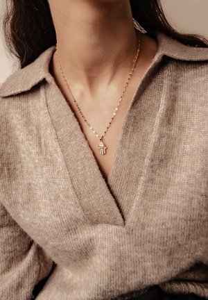 Goldkette mit einem Handanhänger und einem pinken Steindetail. Getragen über einem beigen, gestrickten Pullover mit V-Ausschnitt.