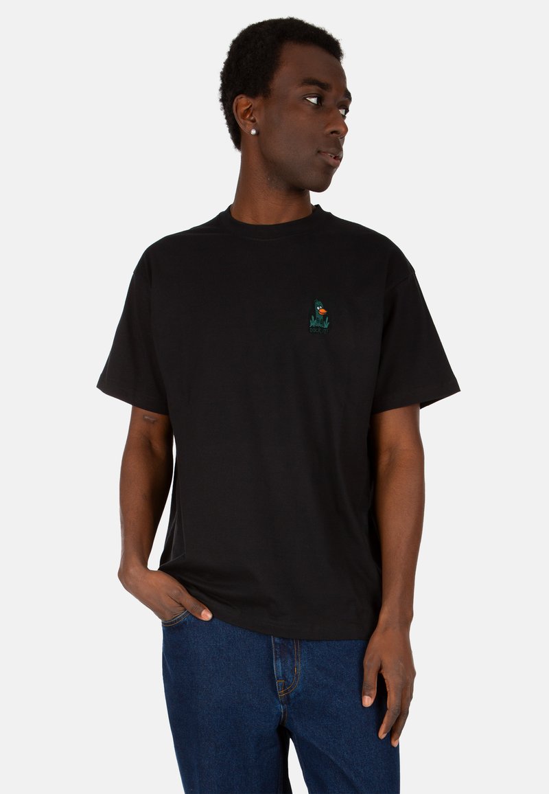 Schwarzes Baumwoll-T-Shirt mit Rundhalsausschnitt, das auf der linken Brustseite ein kleines gesticktes Design eines Charakters in Grün und Orange zeigt.