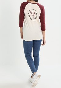 Baseballtröja med trekvartslånga ärmar som har en krämfärgad kropp och burgunderfärgade ärmar. Bakre grafiken har texten "Death Valley Surf Club" och ett hornat skalle.