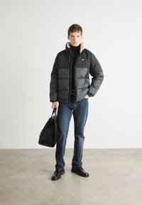 Giacca a vento nera e grigia con colletto crema, abbinata a jeans blu e scarpe nere. Una borsa nera texturizzata è tenuta nella mano destra.