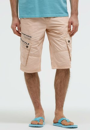 Mann trägt beige Cargo-Shorts mit knopfgedeckten Taschen und blauen Flip-Flops, steht vor einem schlichten hellen Hintergrund.
