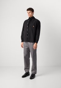Hombre vestido con una camisa negra de botones sobre un jersey de cuello alto negro, pantalones grises y zapatos de vestir negros, de pie contra un fondo blanco.