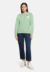 Miętowy sweter z wyszytymi naszywkami, ściągaczami przy mankietach i okrągłym dekoltem, połączony z ciemnoniebieskimi, ściętymi dżinsami i białymi trampkami.