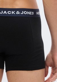 Mees, kes kannab musti Jack & Jones bokserpükse, nähtav vöökohast kuni reie keskosani, käsi lõdvalt kõrval.