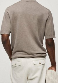 Homme portant une chemise beige texturée à manches courtes rentrée dans un pantalon de couleur claire, tenant un vêtement tricoté dans sa main droite, vue de dos.