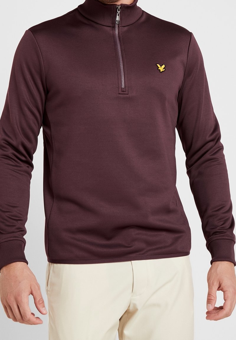 Homme portant un pull à manches longues bordeaux avec une fermeture éclair sur le quart avant et un petit logo d'oiseau jaune sur la poitrine gauche, ainsi qu'un pantalon beige.