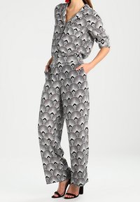 Mustavalkoinen kuviollinen haalari, jossa on kaulus, pitkät hihat ja leveät lahkeet; siinä on koko materiaalin kattava tuuletinsuunnittelu sekä sivutaskut.