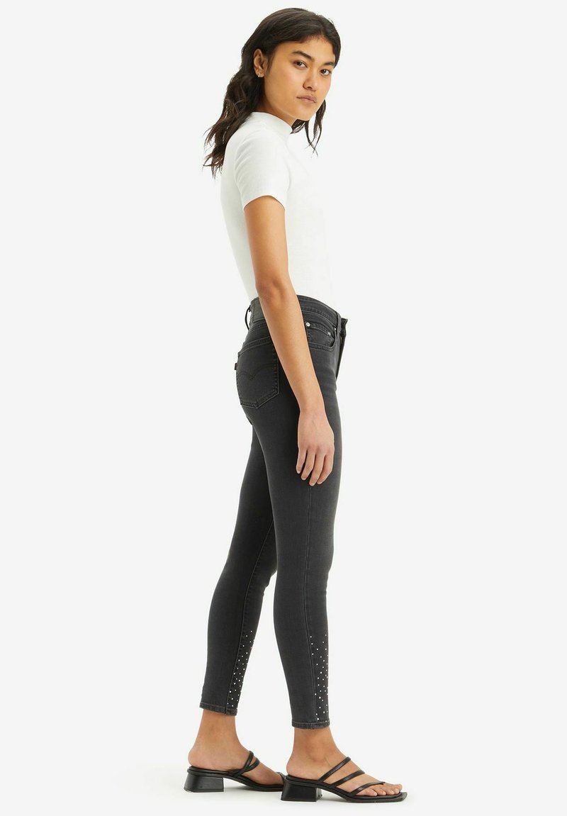 Schwarze Denim-Jeans mit einem schlanken Schnitt, verziert mit Nietenakzenten am Knöchel; kombiniert mit einem weißen, figurbetonten T-Shirt und schwarzen, high-heeled Sandalen.