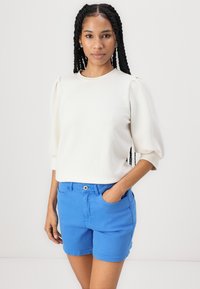 Kermainen collegepaita, jossa on puffihihat, yhdistettynä sinisiin shortseihin. Kangas näyttää sileältä, ja shortseissa on sivutaskut sekä nappisuljenta.