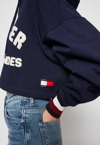 Sweat à capuche court bleu marine avec lettrage texturé blanc ; comporte un patch logo rouge, blanc et bleu ; poignets côtelés avec rayures contrastées.
