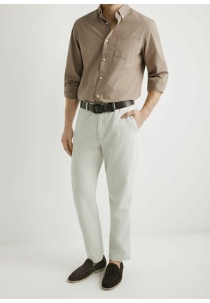 Mężczyzna ubrany w jasnobrązową koszulę z długim rękawem i guzikiem, wpuszczoną w białe spodnie z czarnym paskiem oraz ciemne wsuwane buty.