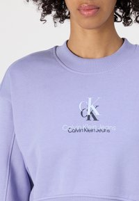 Lila pulóver, bordás nyakkal, középen ezüst színű grafikai logóval: "CK" és "Calvin Klein Jeans" felirat. Puha tapintású.