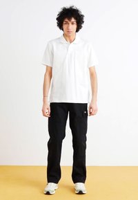 Polo blanc à manches courtes associé à un pantalon cargo noir avec poches latérales, porté avec des baskets blanches. Fond uni.