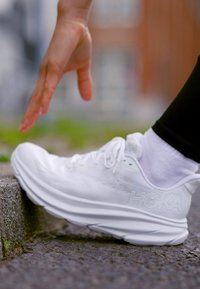 Scarpa da ginnastica bianca con suola spessa, tomaia in tessuto liscio e logo prominente. Mano che si avvicina alla scarpa, su uno sfondo sfocato.