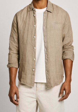 Uomo con camicia beige a maniche lunghe abbottonata sopra una t-shirt bianca e pantaloni chiari con le maniche rimboccate.