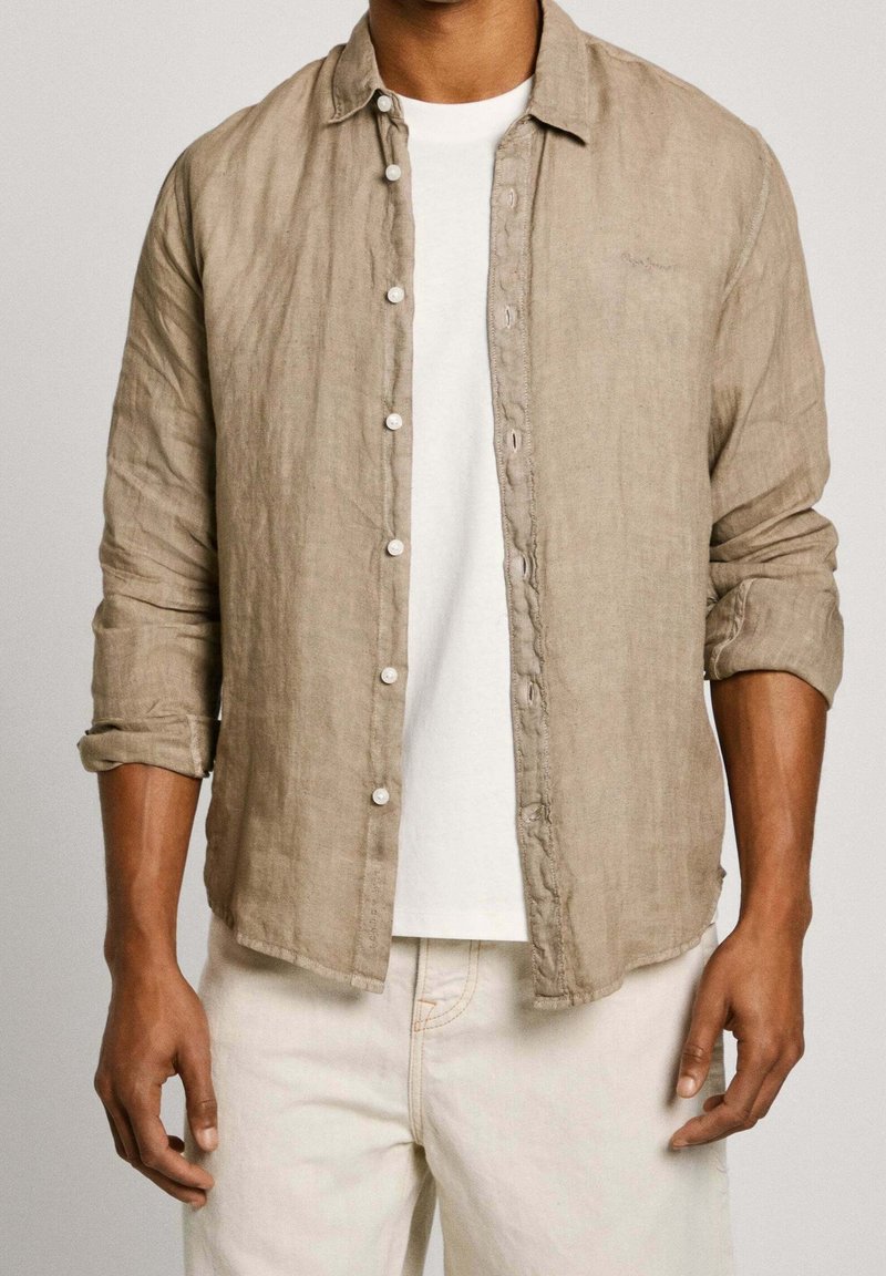 Homme portant une chemise beige à manches longues boutonnée par-dessus un t-shirt blanc et un pantalon clair avec les manches retroussées.