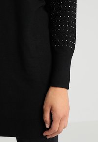 Main d'une femme reposant à côté d'un vêtement noir à manches longues avec de petits clous argentés sur la manche, sur fond clair uni.