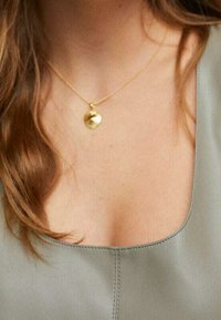 Collier en or en forme de cœur avec un petit pendentif, porté sur une chaîne fine. Le haut présente un décolleté carré de couleur gris clair.