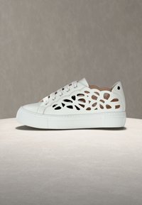 Λευκό αθλητικό παπούτσι με λουλουδάτο μοτίβο κοπής στα πλαϊνά, παχιά σόλα και κορδόνια στο μπροστινό μέρος, τοποθετημένο σε λεία επιφάνεια με φόντο γκρι.