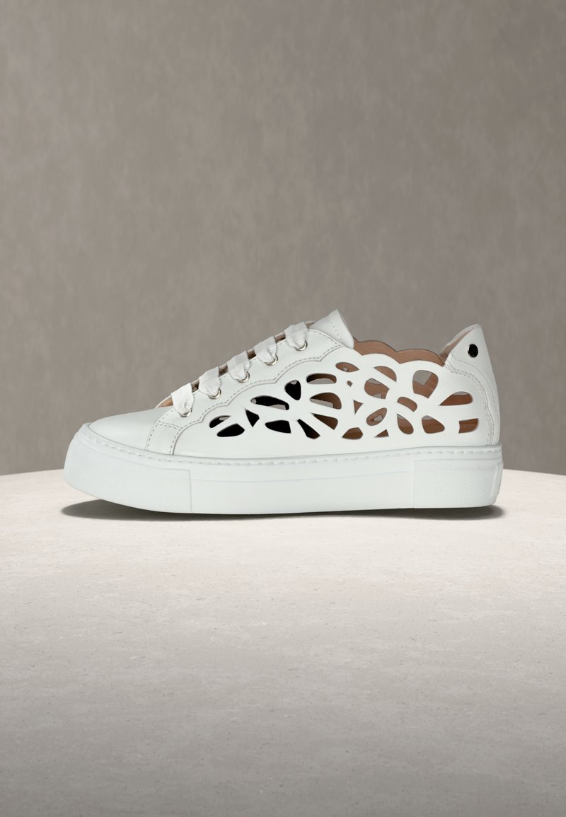 Λευκό αθλητικό παπούτσι με λουλουδάτο μοτίβο κοπής στα πλαϊνά, παχιά σόλα και κορδόνια στο μπροστινό μέρος, τοποθετημένο σε λεία επιφάνεια με φόντο γκρι.