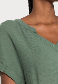 Blusa verde a trama con scollo a V e maniche corte arrotolate. Il tessuto ha un aspetto morbido e stropicciato.