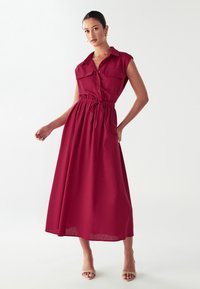 Donna in piedi con un abito midi rosso senza maniche, dotato di un elastico in vita e due tasche sul petto, abbinato a sandali beige con la punta aperta.