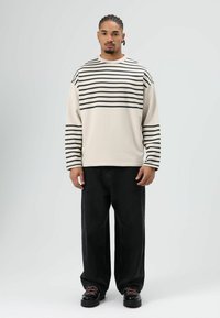 Chemise beige à manches longues avec rayures horizontales noires, associée à un jean noir ample et des chaussures noires avec des accents rouges. Texture lisse.