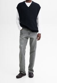 Dunkelblauer ärmelloser Strickpullover über einem weißen Langarmshirt, kombiniert mit hellgrauen Gerade-Passform-Jeans und dunkelbraunen Slippern.