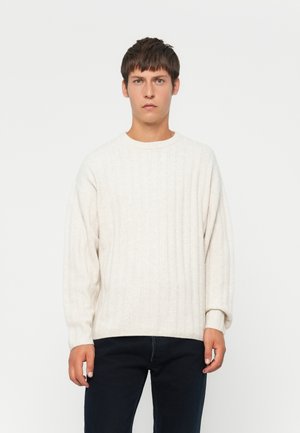 Pull en maille côtelée crème avec un col rond et des épaules tombantes, associé à un jean foncé. Texture douce et coupe décontractée.