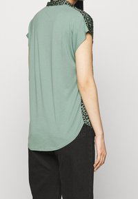 Top verde in mélange di cotone con maniche corte e orlo curvo, caratterizzato da un tessuto a fantasia nero e verde sulle spalle e sui lati.